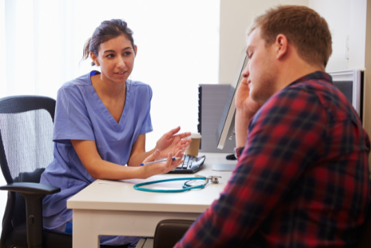 A woman in scrubs speaking to a man in a check shirt with his hand to his head